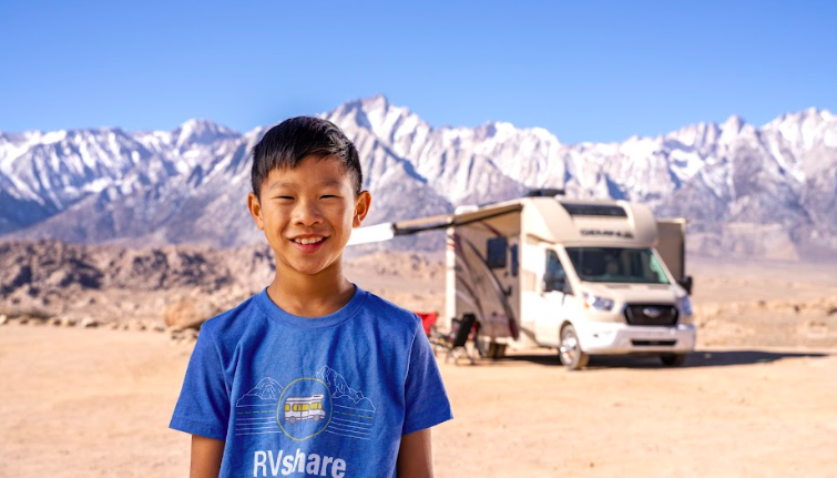 A kid standing in front of a Class C camper by snow capped mountains