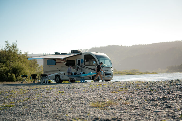 An RV parked next to a river with two men carrying a kayak nearby