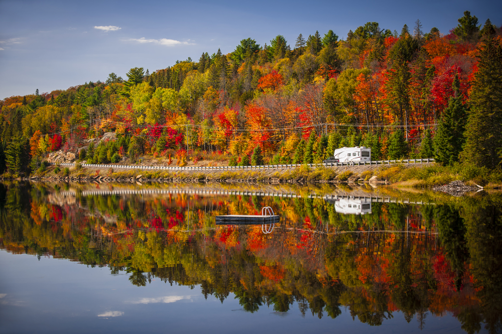 an RV driving next to a lake with fall foliage beyond