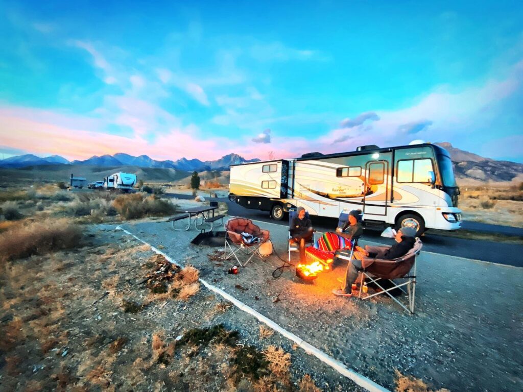 a large RV with people relaxing in front of a campfire