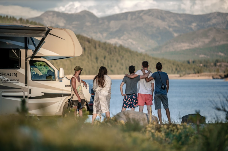 a group of friends next to an RV and in front of a lake