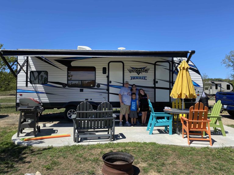 Family of four - mom, dad, son, and daughter - stand outside under the awning of an RV. in the foreground is a table, chairs and umbrella.