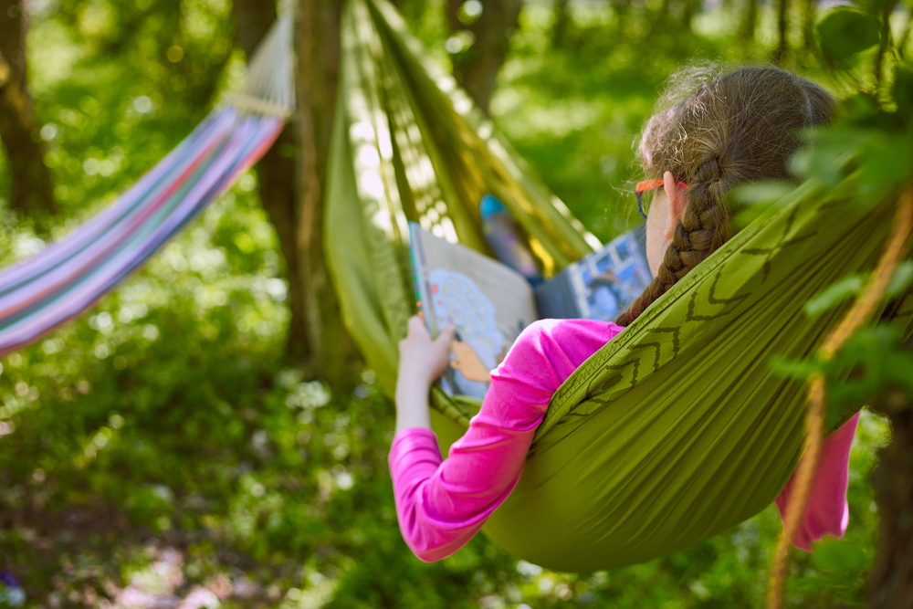 A girl in a hammock reading a book in the woods.