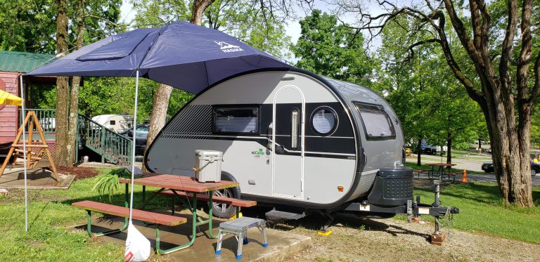 Small light grey teardrop-shaped RV with black horizontal stripe down the center. In front of RV is a wooden picnic table under an umbrella.