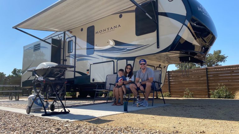 A young family at a campsite, sitting in front of large 'fifth-wheel' RV.