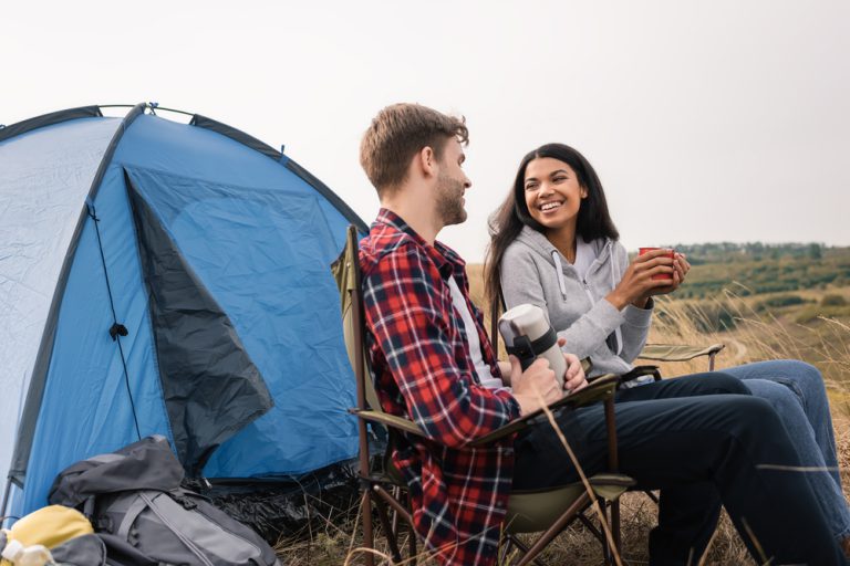 Smiling interracial couple smiling while holding cup and thermos near tent on lawn