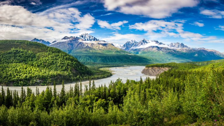 A river runs through a dense, green forest past rocky, snowcapped mountains that are part of the Chugach Alaska Range.