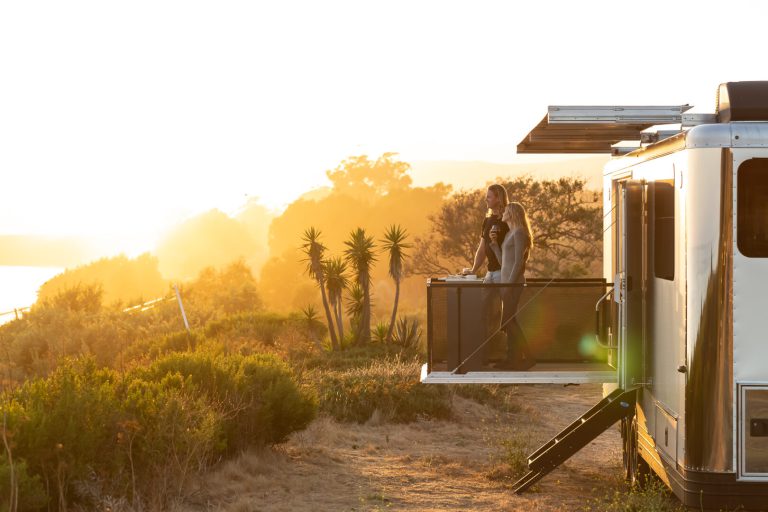 Couple stands on deck off the side of a travel trailer during sunset