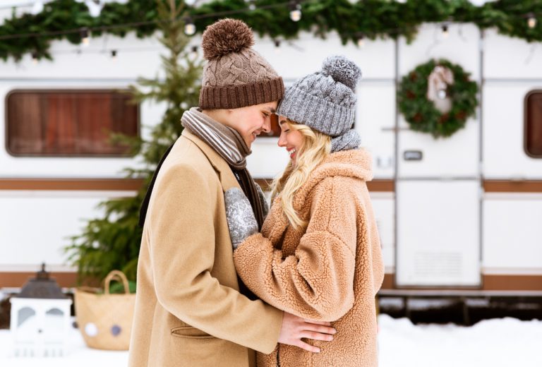 Happy young couple hugging and laughing outdoors in winter, standing near rv trailer motorhome at campsite