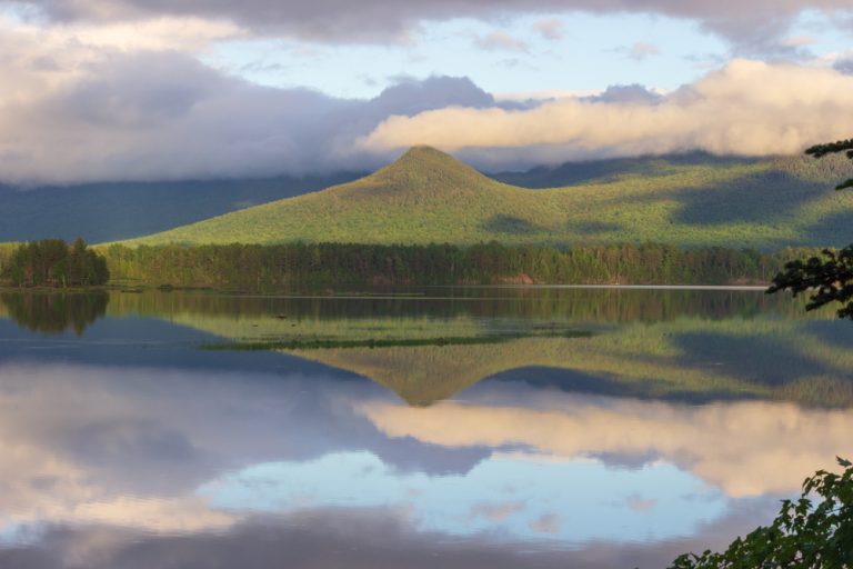 Floating clouds above a green forested mountain peak are reflected on the surface water of Flagstaff Lake in Maine