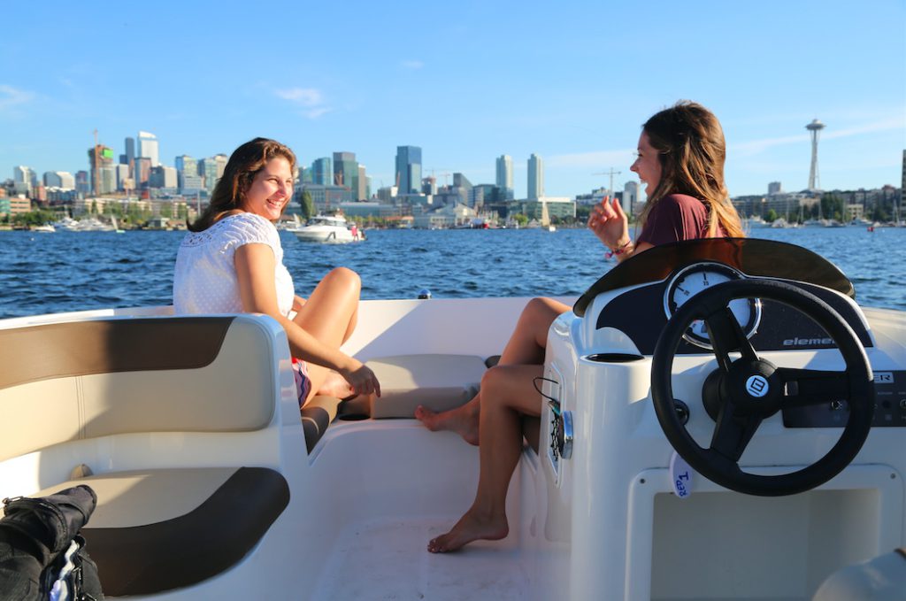Two women in a boat on the water chatting, city skyline behind them