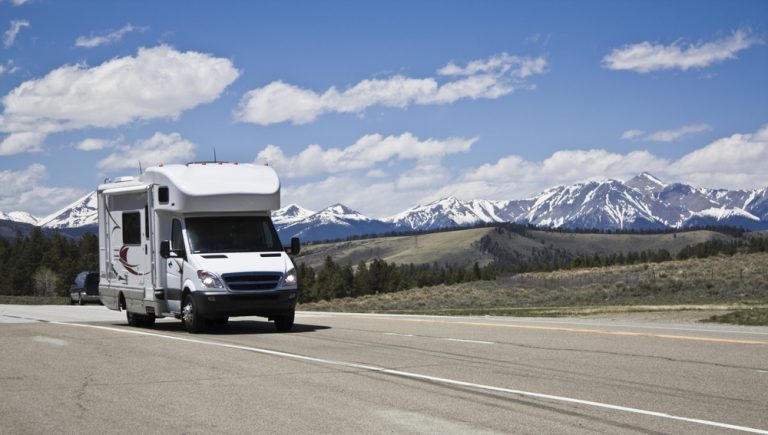 RV traveling on a three-lane highway with mountains, rolling hills, and blue sky with puffy, white clouds in the background