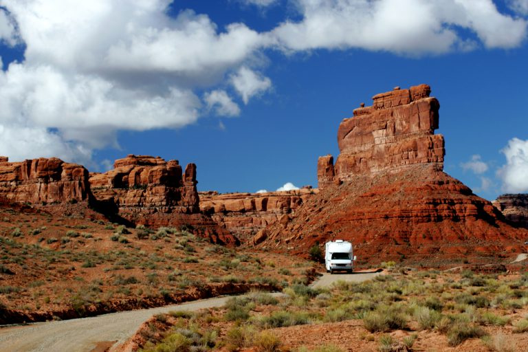 A white RV drives on a road next to the tall, rocky monuments of Utah's Valley of the Gods
