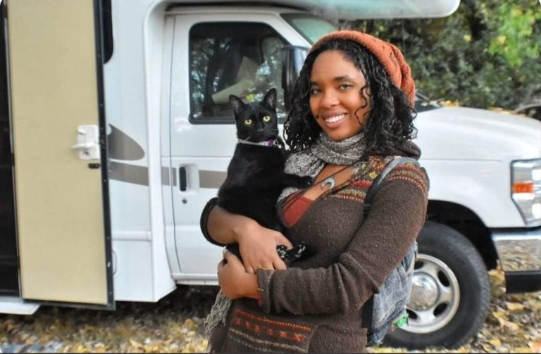 Young woman stands in front of her Class C RV holding her black cat