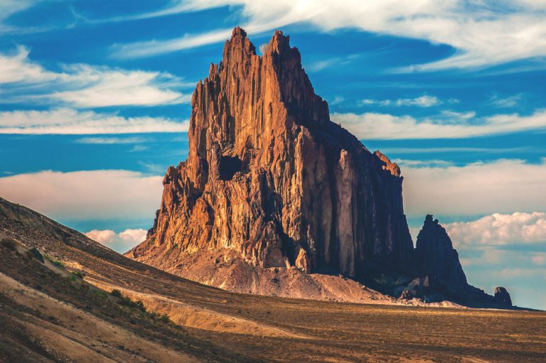 A tall, jagged red-rock butte stands alone in an empty desert under a blue sky.