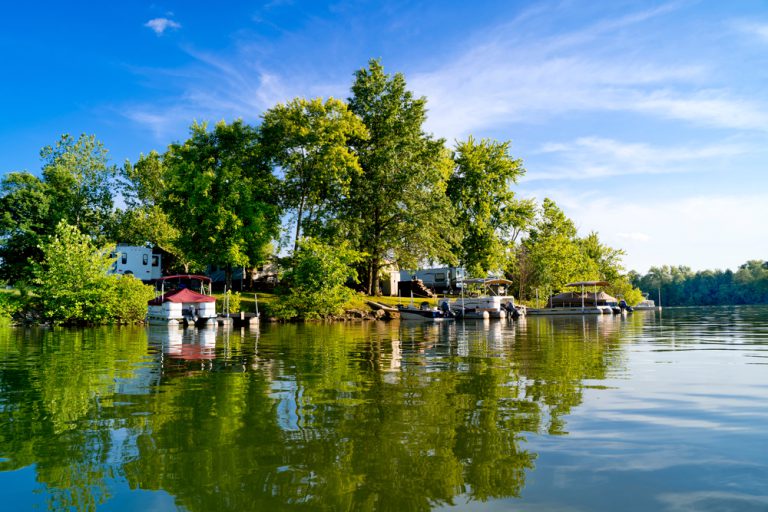 Several RVs and boats sit along the shore of a lake lined with trees with a partly cloudy blue sky overhead.
