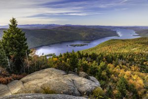 A rocky overlook on a mountain looks out towards a green and yellow forest and shining blue lake.
