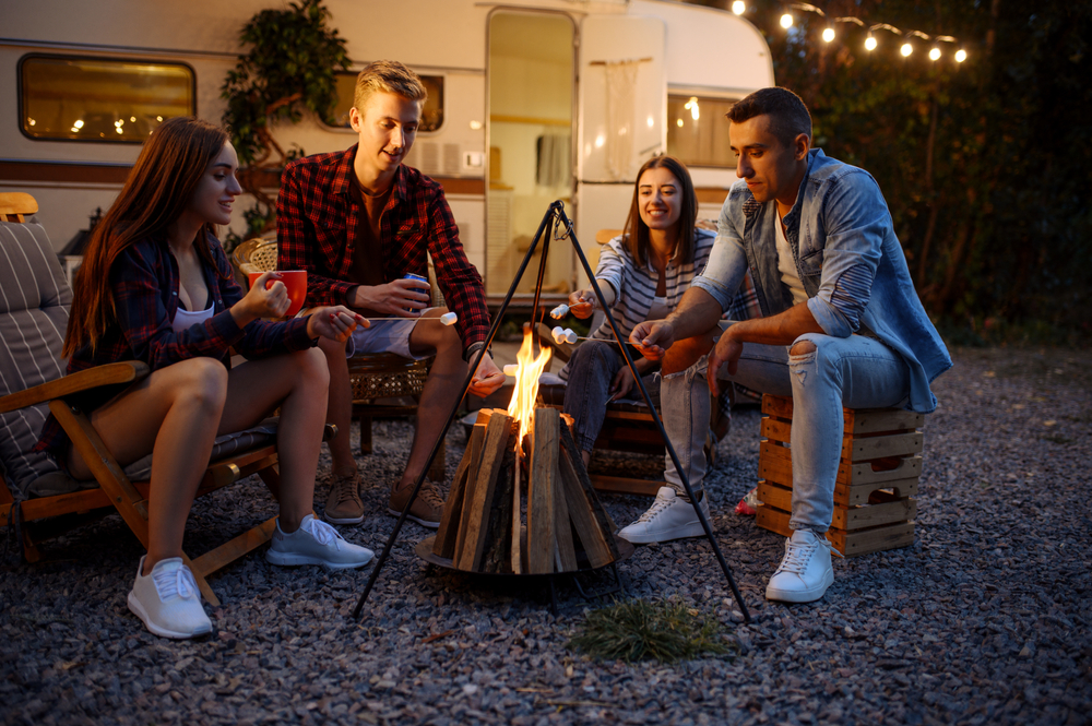 Four friends sit around a campfire outside a parked trailer