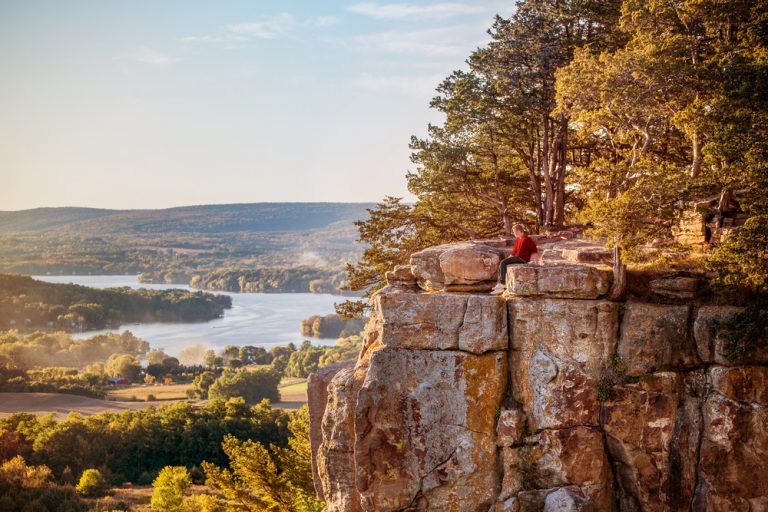 A person sits on a rocky outcropping, high above fields of trees and a lake.