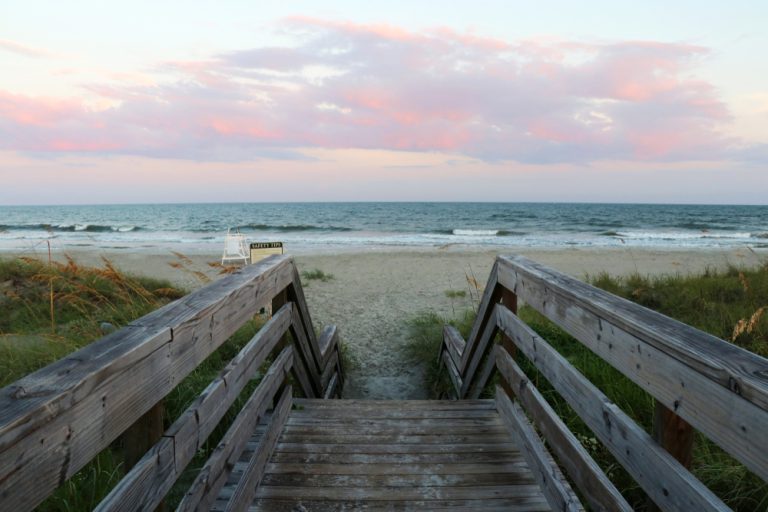 A wooden boardwalk with railings leads to an open, empty beach. The sun is still pink from the sunrise.