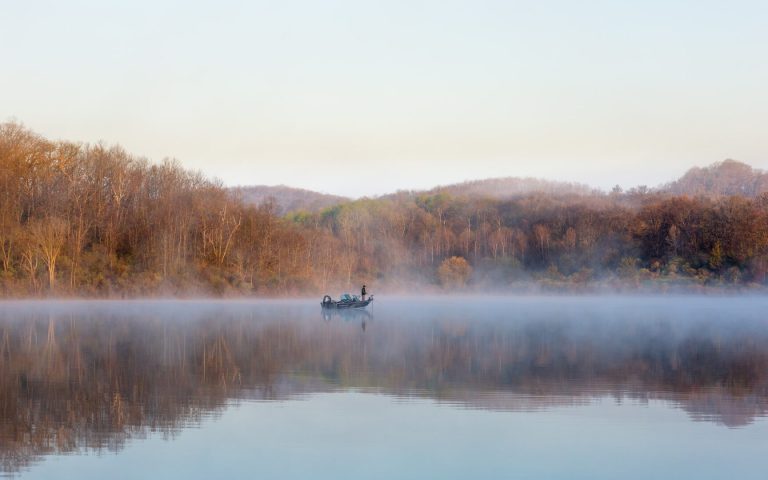 A man stands on a boat in the middle of a calm lake, as fog lifts from the surface of the water. The lake is surrounded by autumn-colored trees.