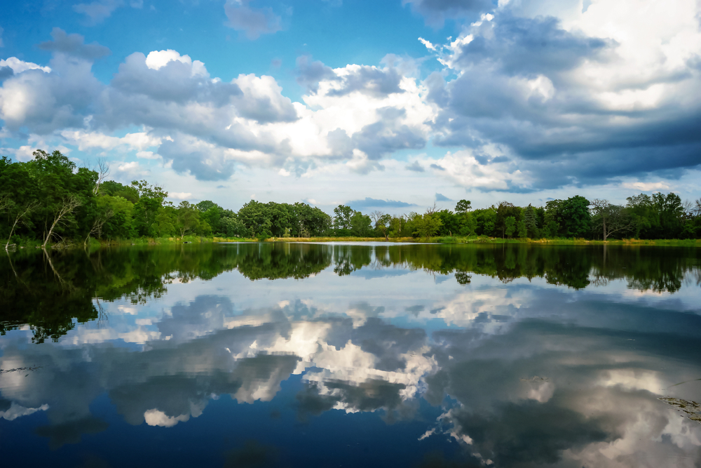 A calm lake reflects fluffy clouds and the surrounding green forest.