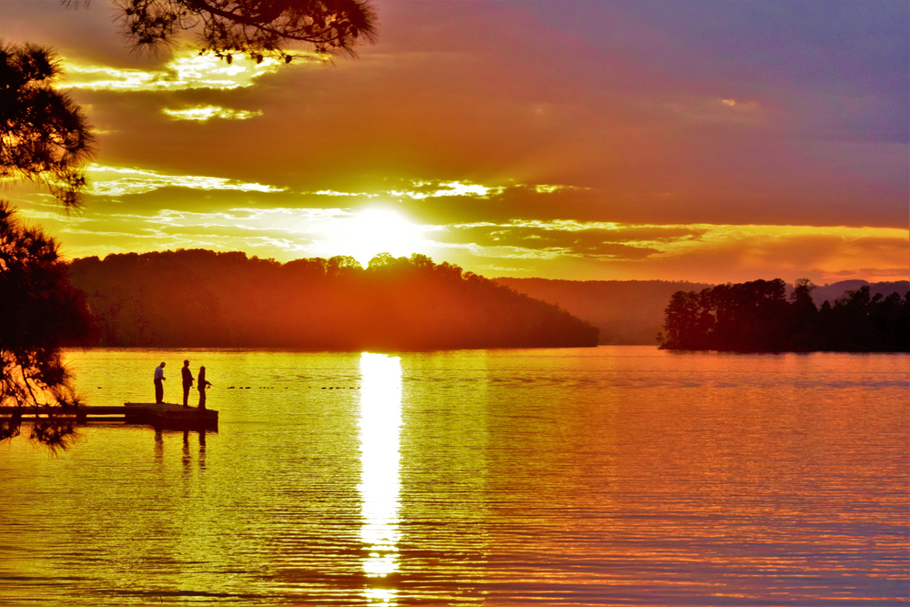Three people fish off a dock in a lake, turned gold by the rising sun.
