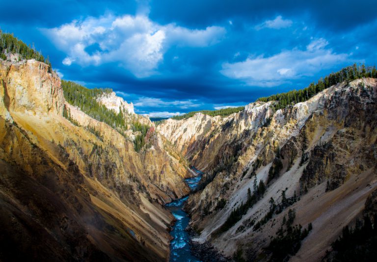 A river flows through the middle of a canyon with steep walls, under a blue, cloudy sky.