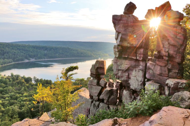 The sun shines through a unique rock formation atop a rocky cliff, looking out over a forest and lake.