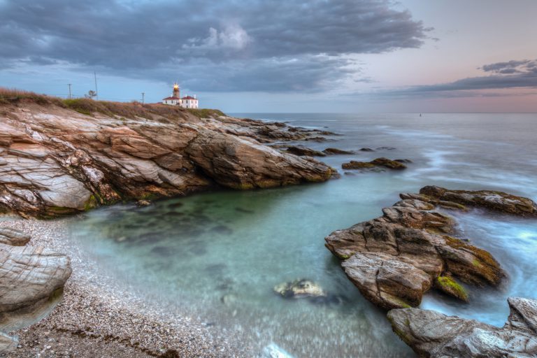 Waves roll towards a rocky beach shore, a lighthouse sitting on the beach in the distance.