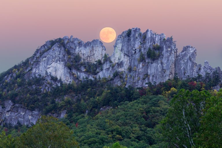 A supermoon rises over the dip in the middle of a unique rocky mountain, Seneca Rocks. The foothills are covered in green forests.