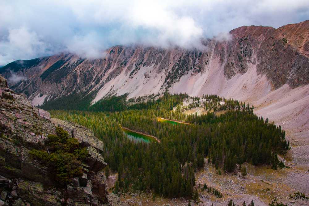 A thin lake in the middle of a verdant green forest rests at the bottom of a huge canyon under an overcast sky.