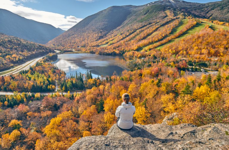 A woman sits on a granite cliff overlooking a forest in autumn covers surrounding a lake at the foothills of a mountain.