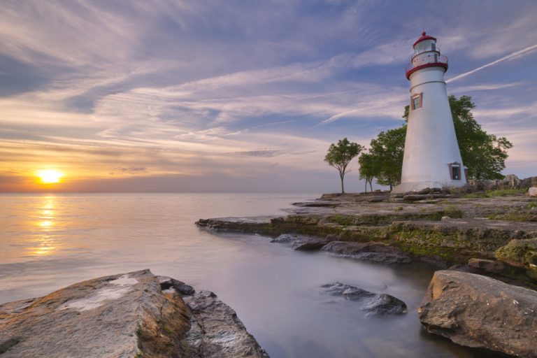 A white lighthouse sits on the shore of a lake as the sun sets in the distance.