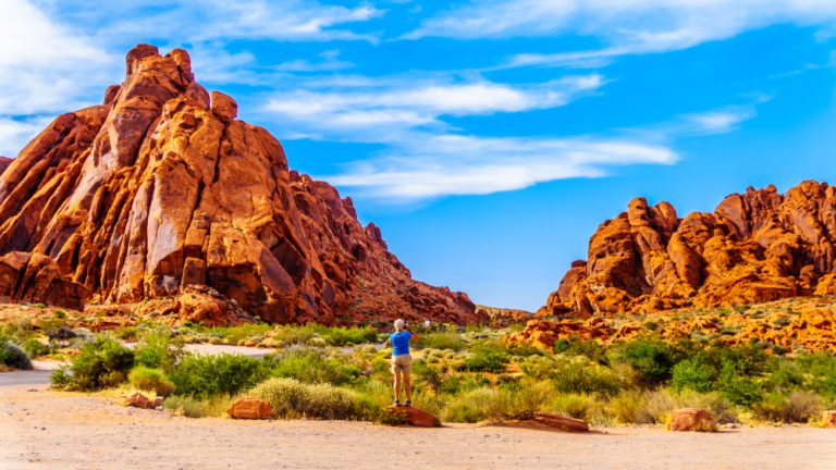 A person in a blue jacket and khaki shorts stands on the side of a dirt path to take a photo of two unique red-orange rock formations under a bright blue sky.