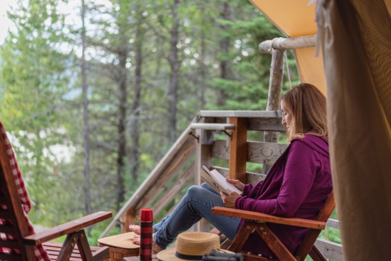 A woman in a purple jacket and demin jeans sits in a wooden porch chair reading a book, surrounded by a forest of thin trees.
