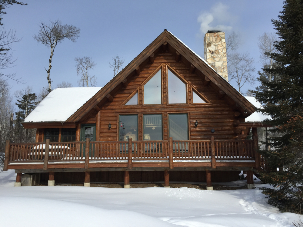 A wooden cabin with large front windows and a smoky chimney sits in a snow-covered clearing surrounded by trees.