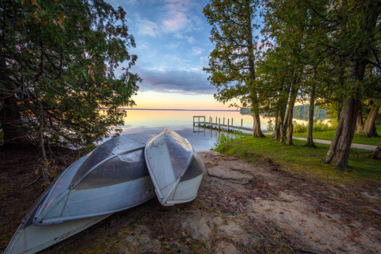 Three aluminum canoes rest on the shore of a lake, off the side of a dirt path, surrounded by dense trees.