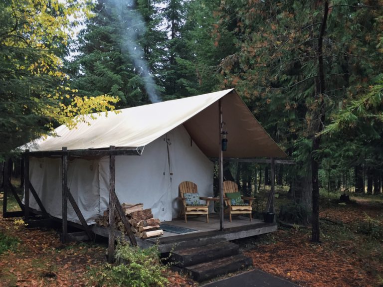 A luxury tent sits in the middle of a forest. There are two chairs and a woodpile on its porch, and smoke rises from the back.