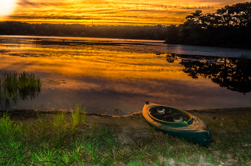A kayak sits on the grassy shore of a lake colored orange and reflecting the clouds at sunset.