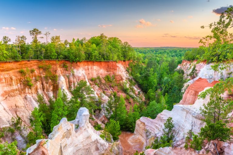 A canyon made up of red and white rock formations, with verdant green trees growing in the base and around the rim of the formations. The sunset tinges the sky pink and orange in the distance.