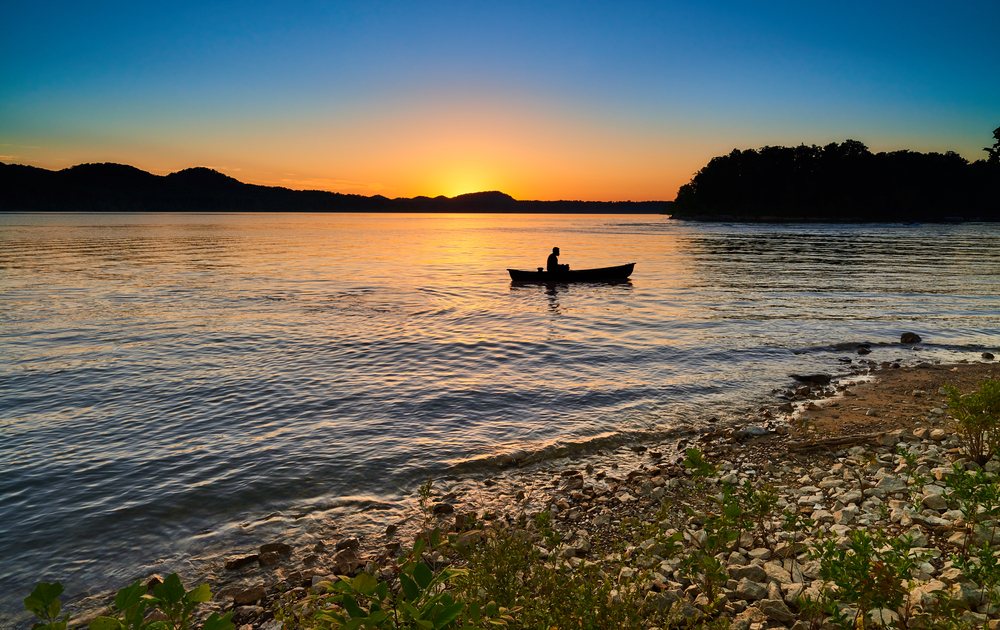 A canoeist on a lake is silhouetted by the setting sun, turning the sky orange. The lake shore is rocky and covered with grass.