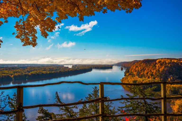 An overlook with a wooden guardrail stands above the Mississippi river, the trees turning autumn colors.
