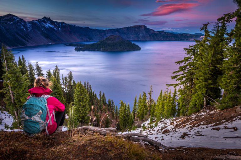 A hiker with a pink jacket and blue backpack sits on the ground, looking out over a large mountain lake with an island jutting out of the water. The sunset paints the sky and water pink and purple.