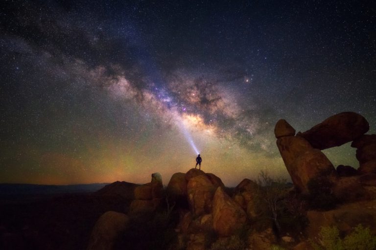 A person standing on a rocky hill looks up at the night sky, illuminated by a headlight, which is a stunning view of the Milky Way.