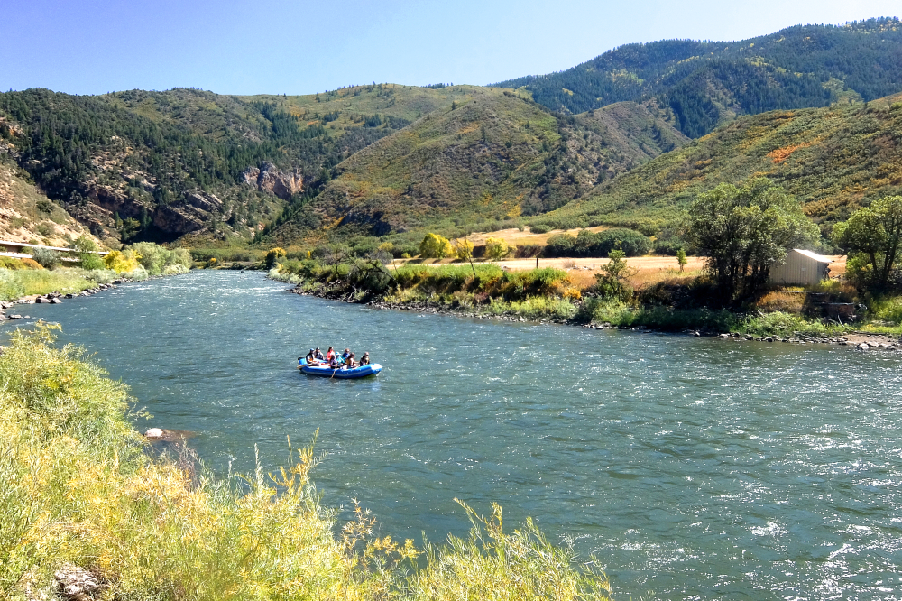 A group of people on a raft in a wide river, a cabin a little ways back from the shore.