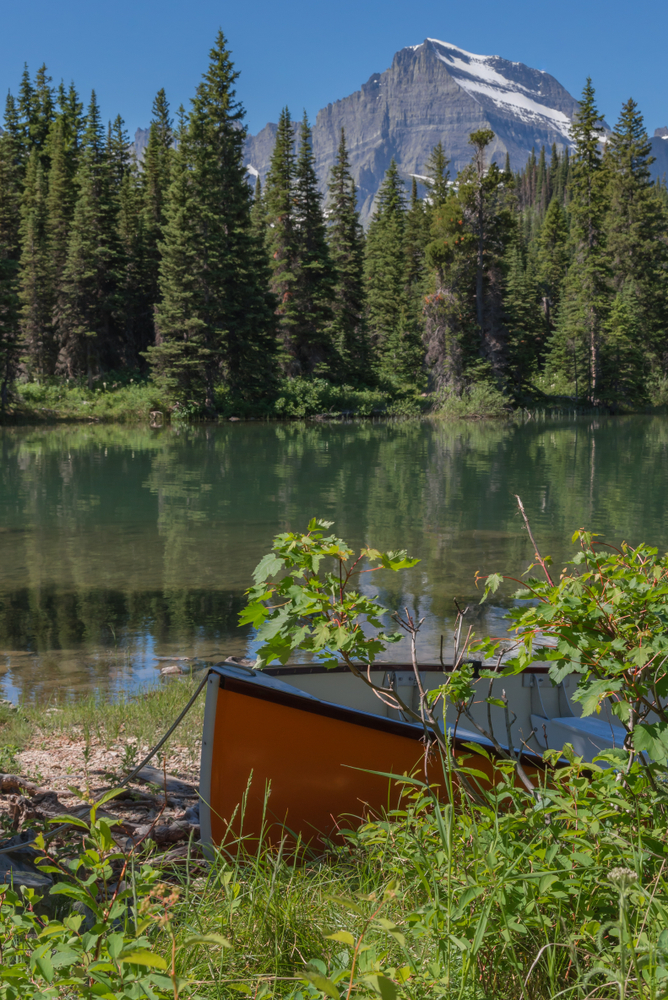 Orange canoe hidden in the brush along a mirror-still forested lake with a mountain rising in the background along the Swiftcurrent trail in Glacier National Park in the American state of Montana.