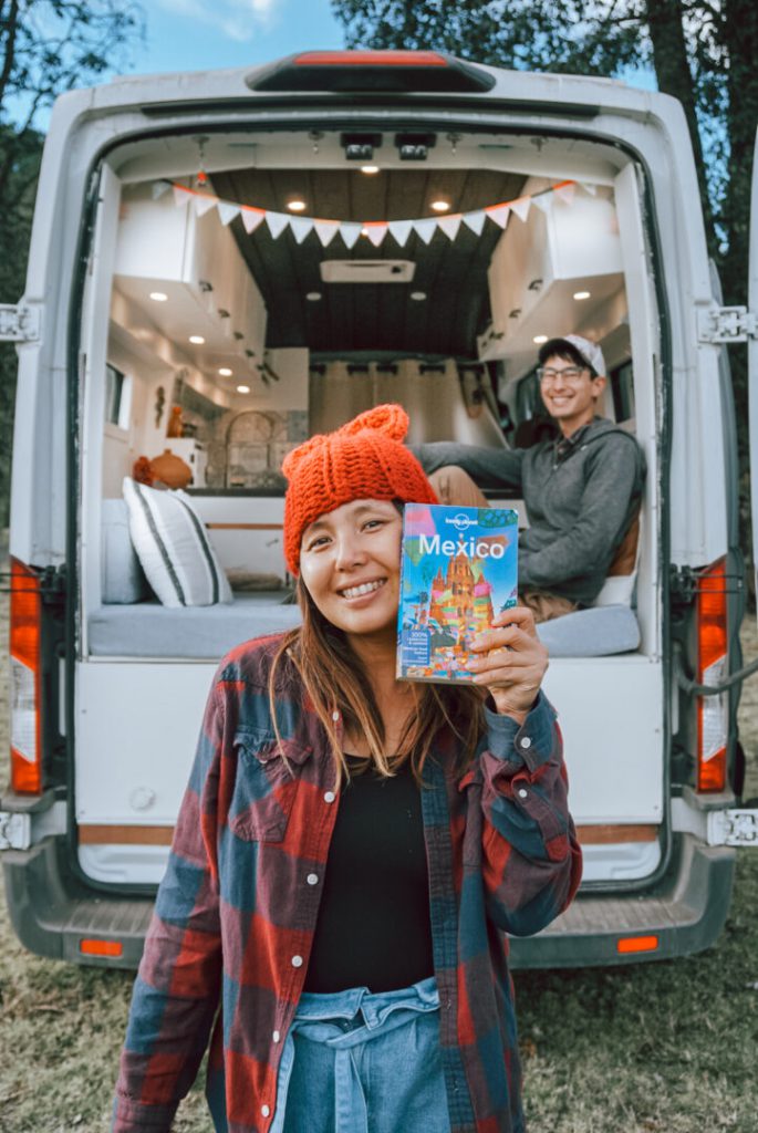 Woman holds a travel guide to Mexico with her campervan and boyfriend in the background