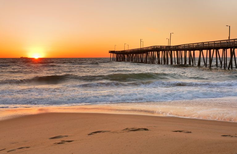 A long wooden pier stands above a ocean, waves crashing toward the shore as the sun sets over the horizon.
