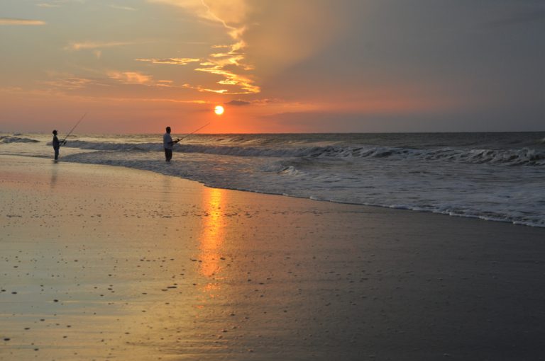 Silhouette of a man and a boy fishing in the ocean against an orange, yellow, and grey sunrise.